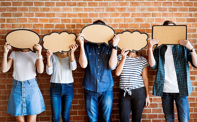 Line of people standing against a brick wall holding large thought bubbles over their faces.