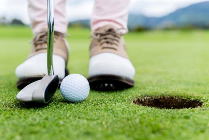 Close-up shot of golf shoes, the base of a golf club, and a golf ball on green grass.