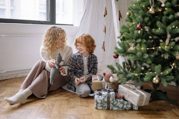 A mother and child sitting next to a pile of gifts under a Christmas tree.