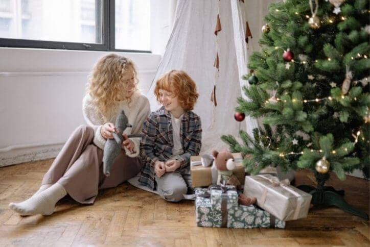 A mother and child sitting next to a pile of gifts under a Christmas tree.