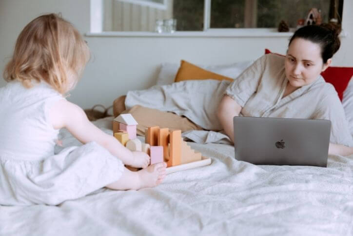 A toddler plays with wooden blocks nearby a woman on her laptop.