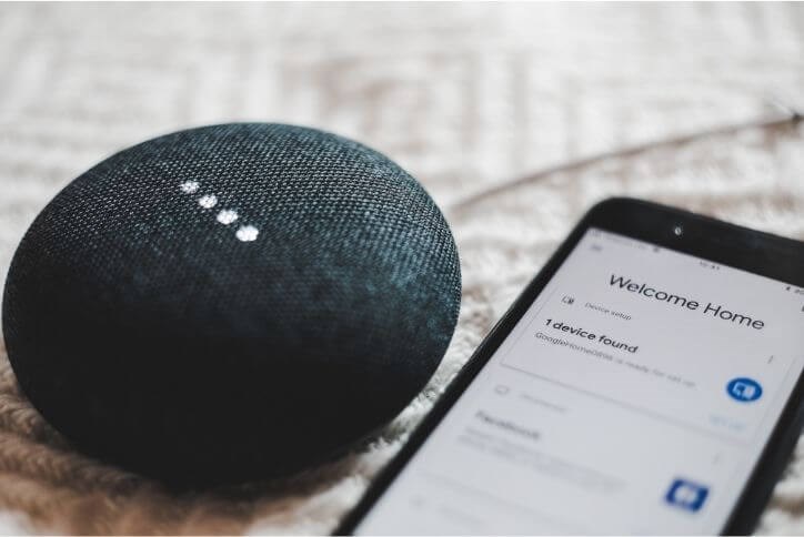 Alt text: Round gray smart speaker next to a smartphone with welcome home on the screen.