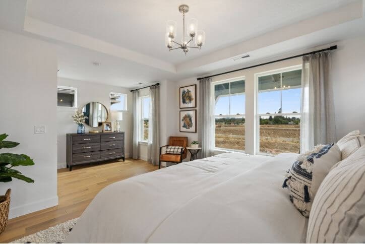 A bedroom with large windows, white linens, and a simple wood dresser.