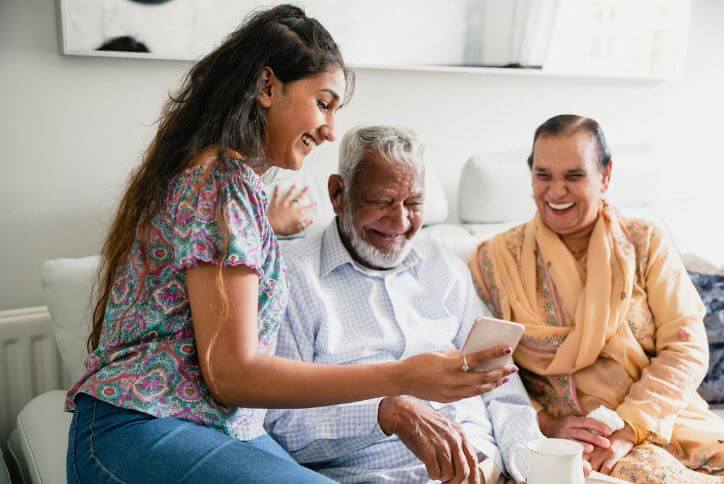 An older Indian man, woman, and young woman laugh while looking at a mobile phone.
