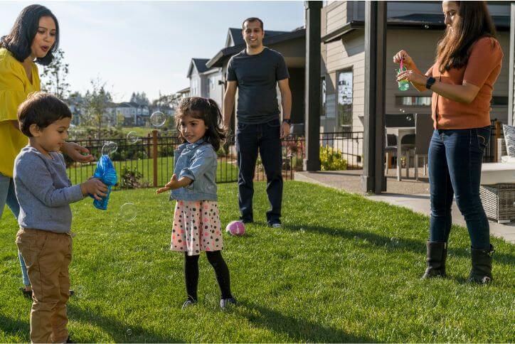 A family with two small children stands in a grassy backyard, blowing soap bubbles.