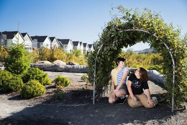 Two teenage sisters smile at each other under an arch made of leafy green branches in Reed’s Crossing.