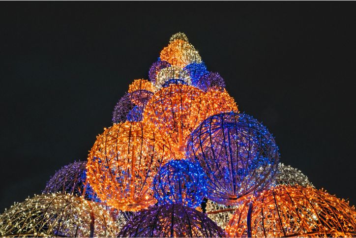 A holiday light display features large balls wrapped with lights stacked to form a pyramid.