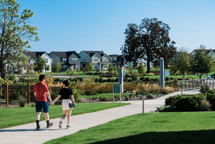 A young couple walks hand-in-hand along a path in Reed’s Crossing, with Tamarack Park visible in the background.