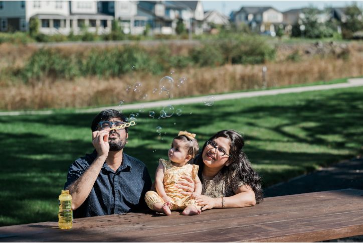 A baby in a yellow dress sits on a picnic table as her parents blow soap bubbles around her at Dobbin Park in Hillsboro, OR.