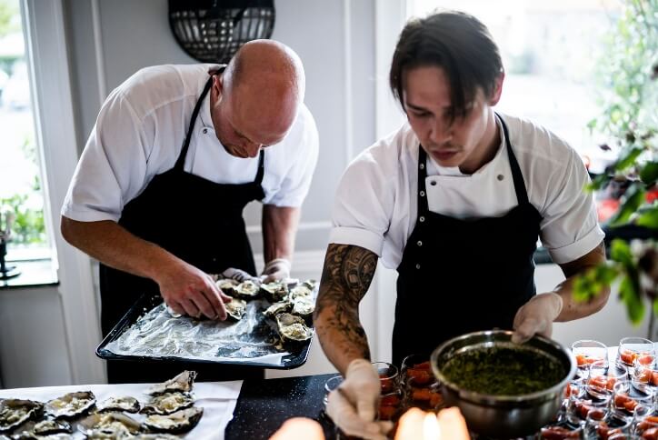 Two men in black aprons prepare food in a brightly lit kitchen.