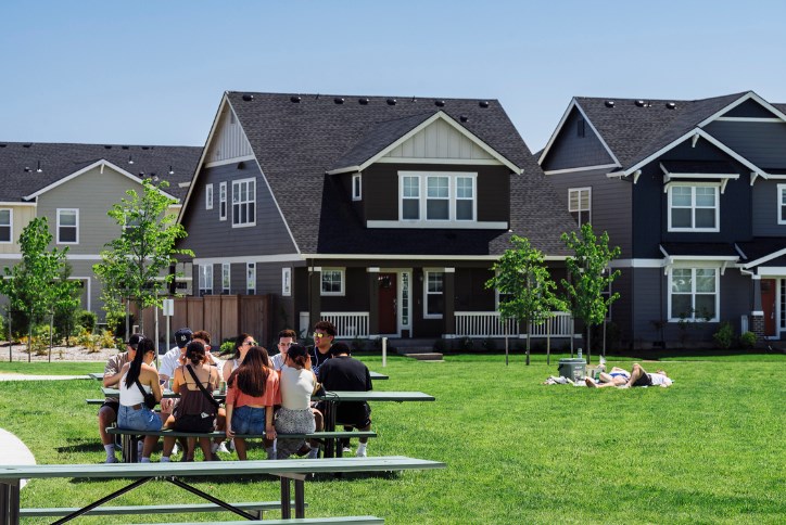 A group of young people sit at a picnic table in Reed’s Crossing Park while a couple lies sun tanning in the grass nearby.