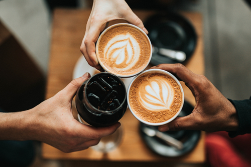Overhead shot of three people toasting with coffee drinks above a wooden table.